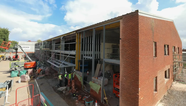 OMX construction site with a partially built brick building, scaffolding, and workers in safety gear standing near the structure. The building’s frame and insulation are visible