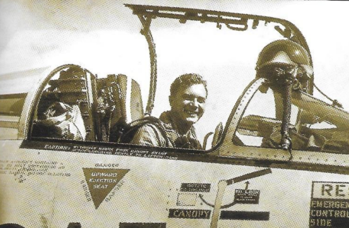 Black and white newspaper photo of a smiling man sitting in the cockpit of a fighter jet