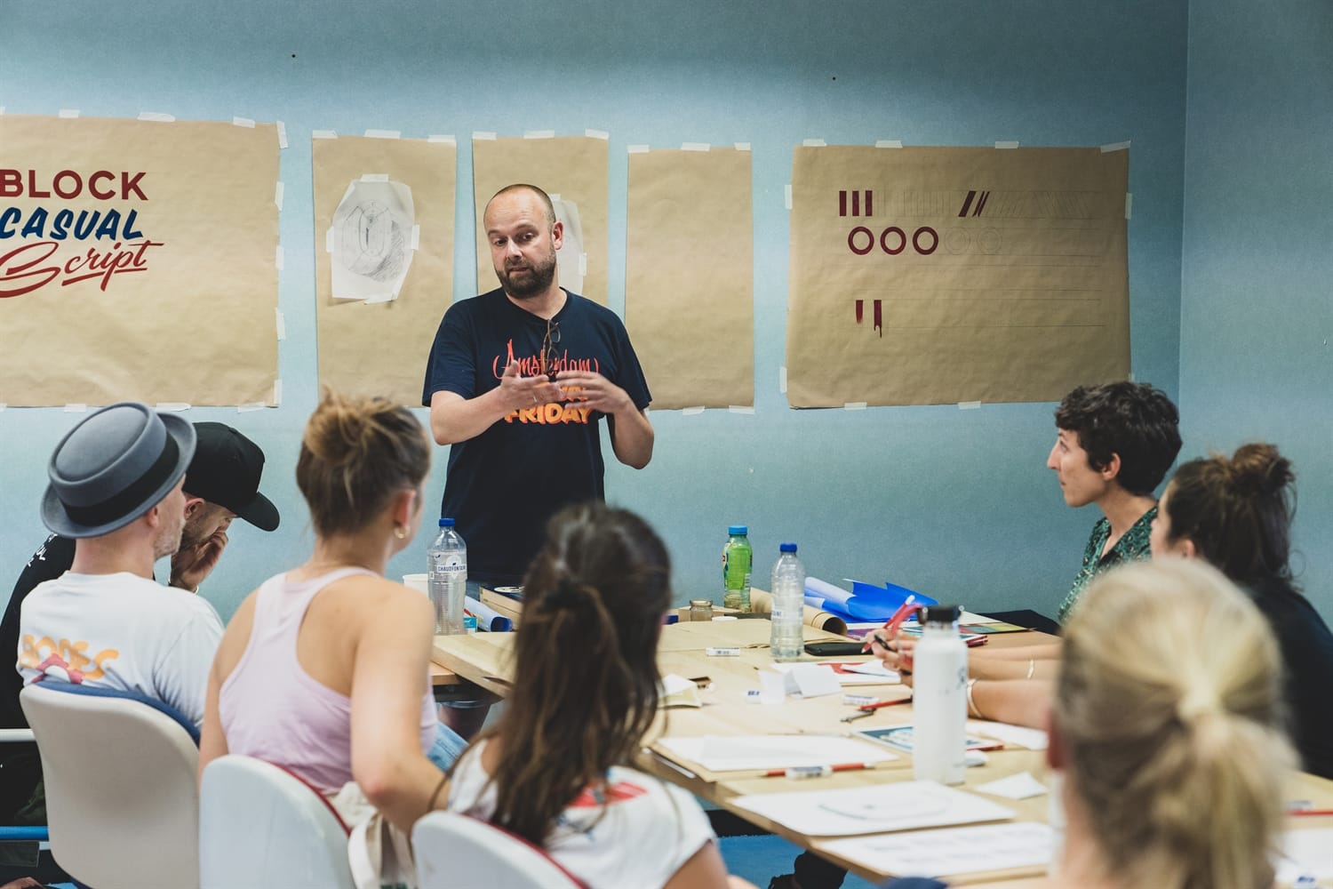 Students at a table listening to an instructor talking about painting letters and lettering effects.