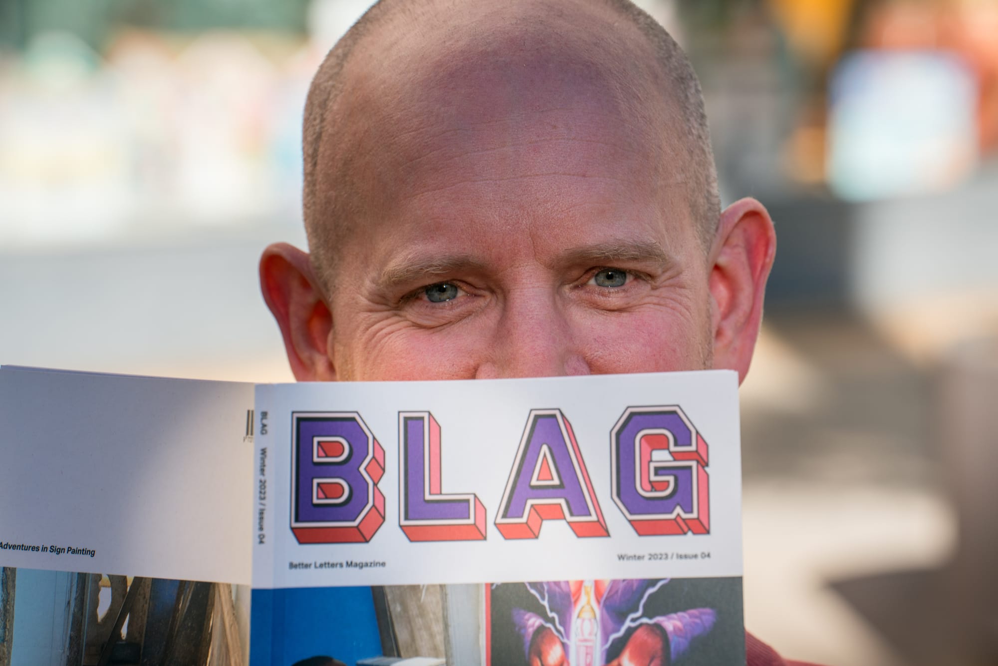 Top half of a man's face smiling while the lower half is obscured by a magazine called BLAG.