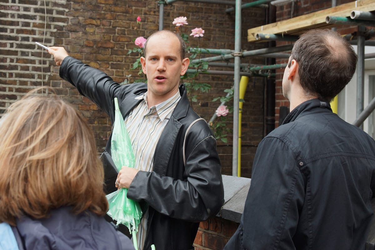 Man in leather jacket holding a green plastic umbrella and pointing at the wall behind him while others look on.