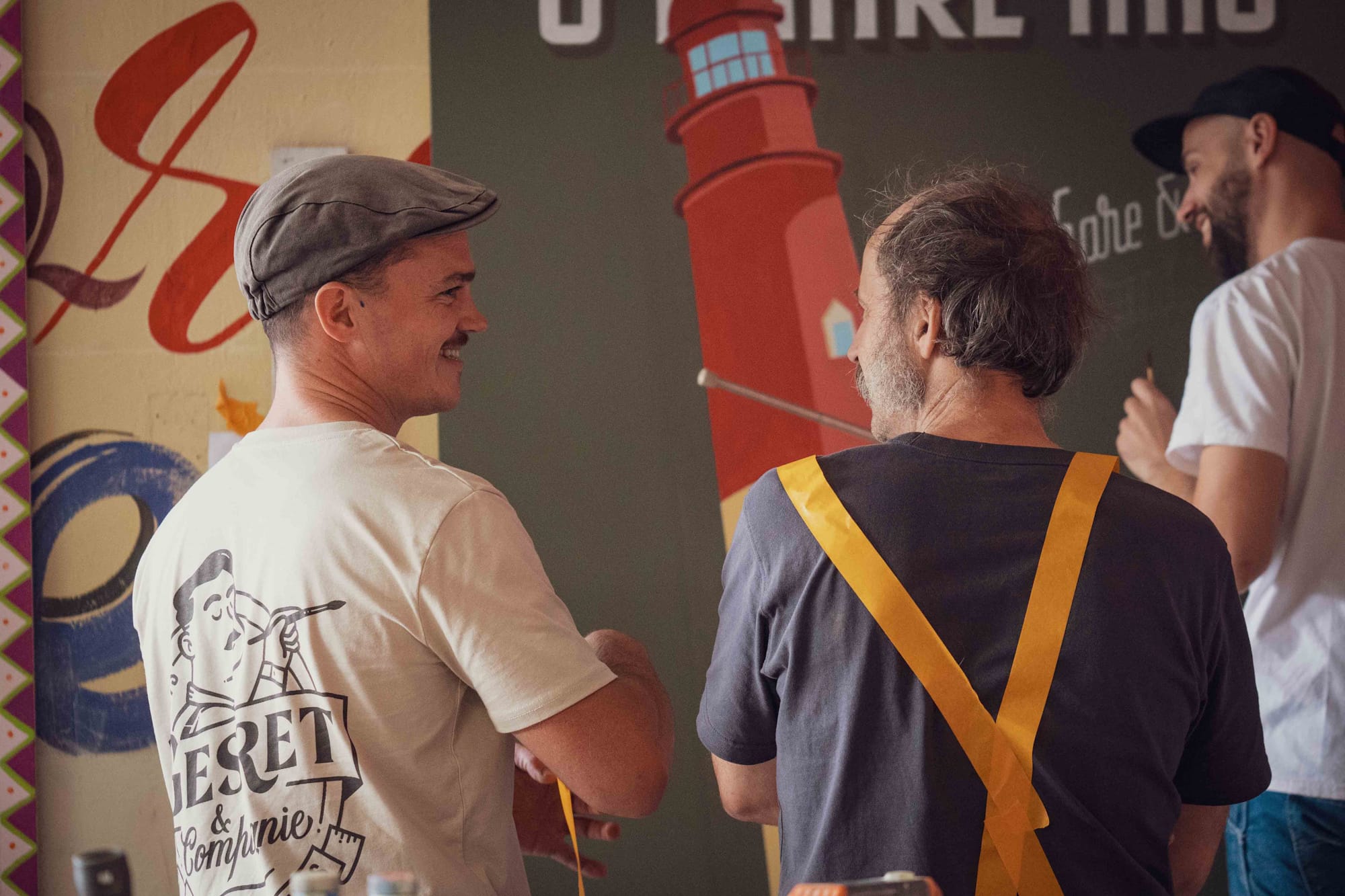 View from behind of two men looking at each other as they talk. In the background another man, slightly out of focus, is pausing from painting a sign, looking to his left and smiling.