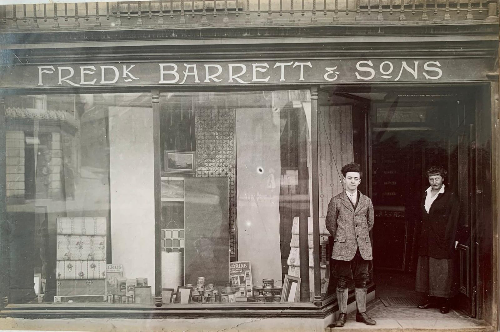 Smartly dressed boy standing in the entranceway to a shop, above which is signwritten 'Fred'k Barratt & Sons'.