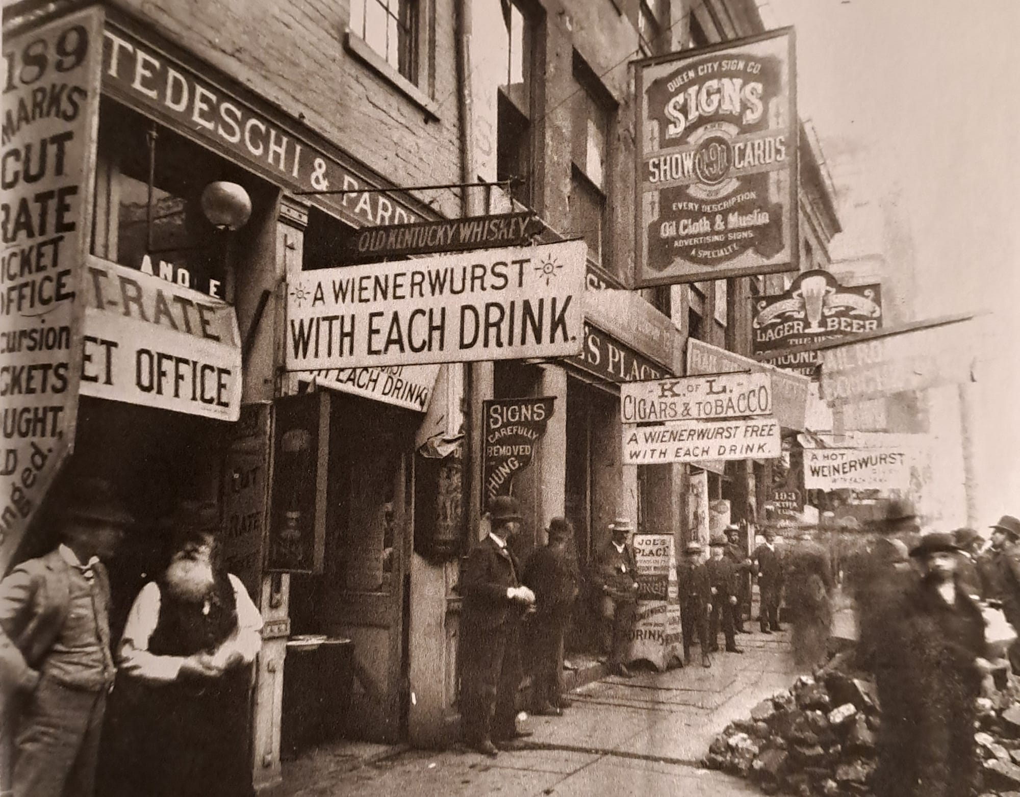 Archival photo showing a street scene filled with traders and a riot of signs protruding from every building. One of these is advertising the "Queen City Sign Company" and their various sign services.