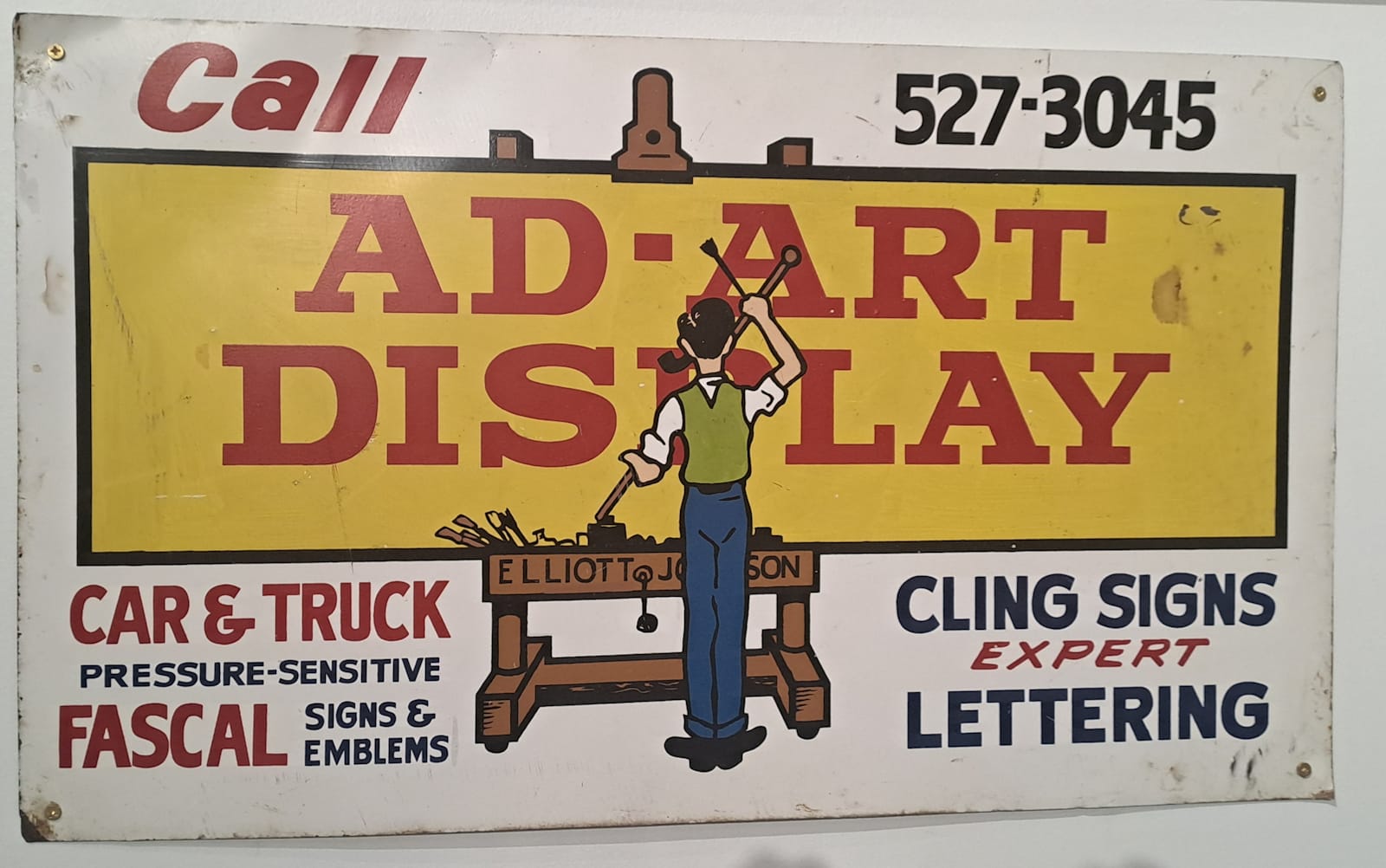 Hand-painted sign set on a pictorial of a man painting a large sign on an easel. The sign on the easel reads "Ad-Art Display", and the easel is lettered with "Elliott & Johnson". The secondary copy set above and below this pictorial element gives the various sign types on offer and the phone number.