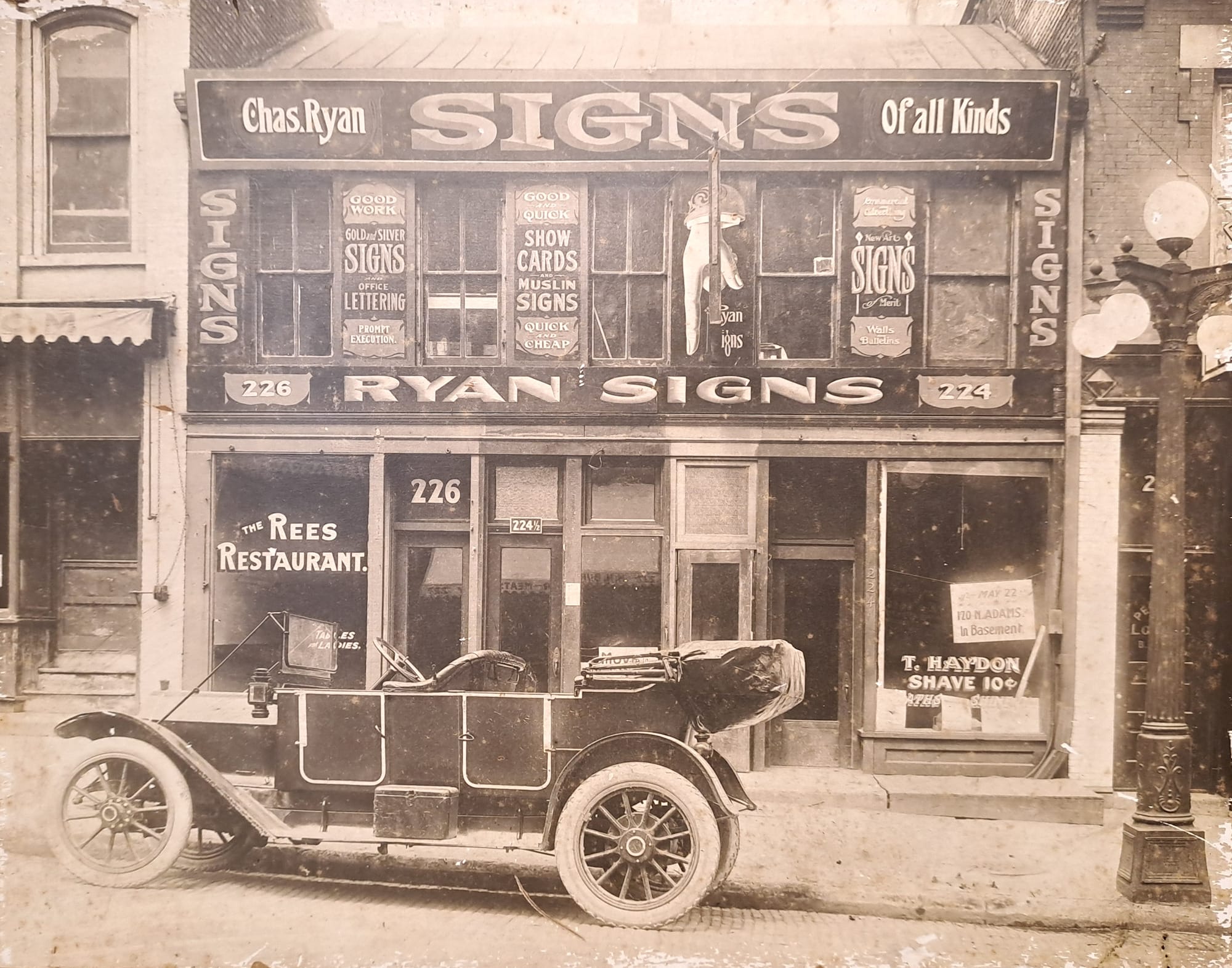 Old car in front of a terraced shop whose first floor is emblazoned with signs advertising the Ryan Signs business.
