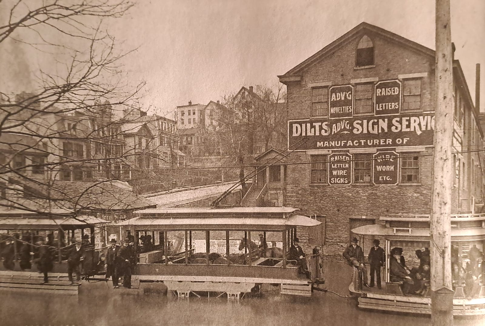 A streetcar with passengers passing along a flooded road. Behind this is an industrial building with painted signs on the walls advertising the "Dilts Advg. Sign Service".
