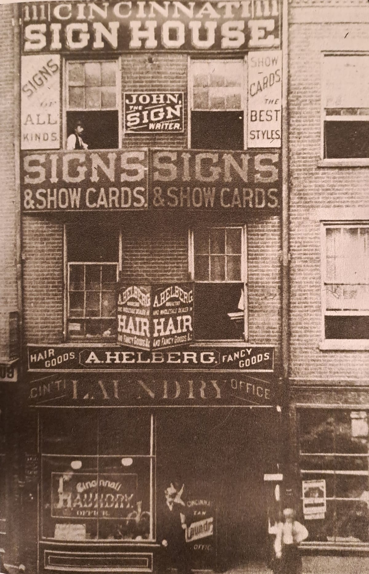Archival photo that shows a tall terraced shop above which there are a host of signs advertising the "Cincinnati Sign House" and "John the Sign Writer".