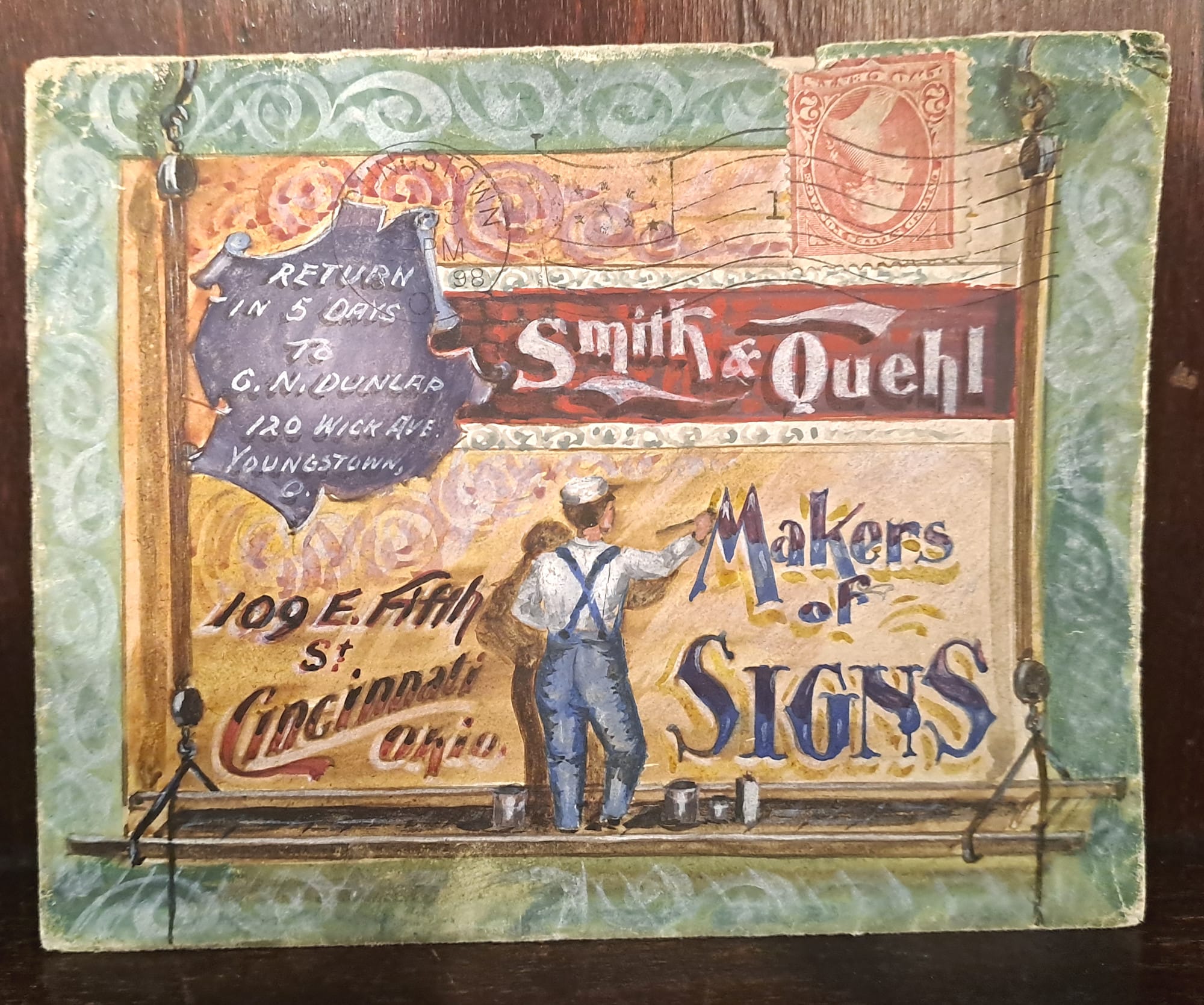 Hand-drawn envelope with a postmarked stamp. Every space is filled with a drawing of a man on a hanging platform painting a billboard. The billboard comprises a design in the form of an advertisement for "Smith & Quehl, Makers of Signs" that doubles as the address for that firm.