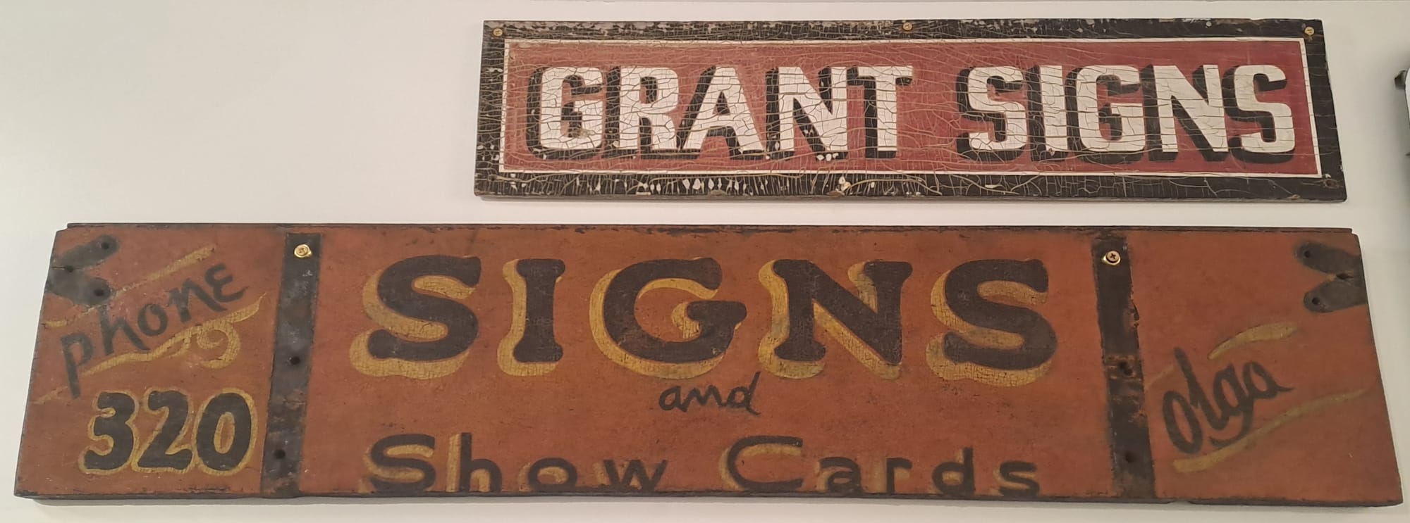 Two wide-format hand-painted signs on a wall. The one above simple says "Grant Signs", while the quite roughly lettered one below has "Signs and Show Cards" in a central panel with "Phone 320" on the left, and just "Olga" on the right.