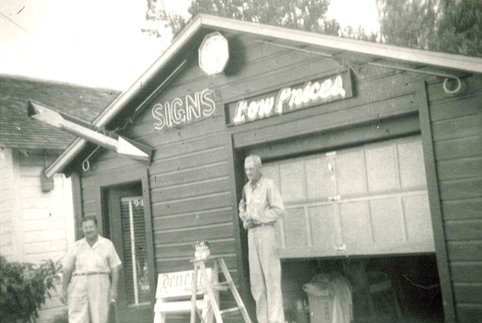 A blurry black and white photo of two men outside a garage building that is crudely adorned with a big arrow, a clock, a neon sign that says "Signs" and another sign that says "Low Prices".