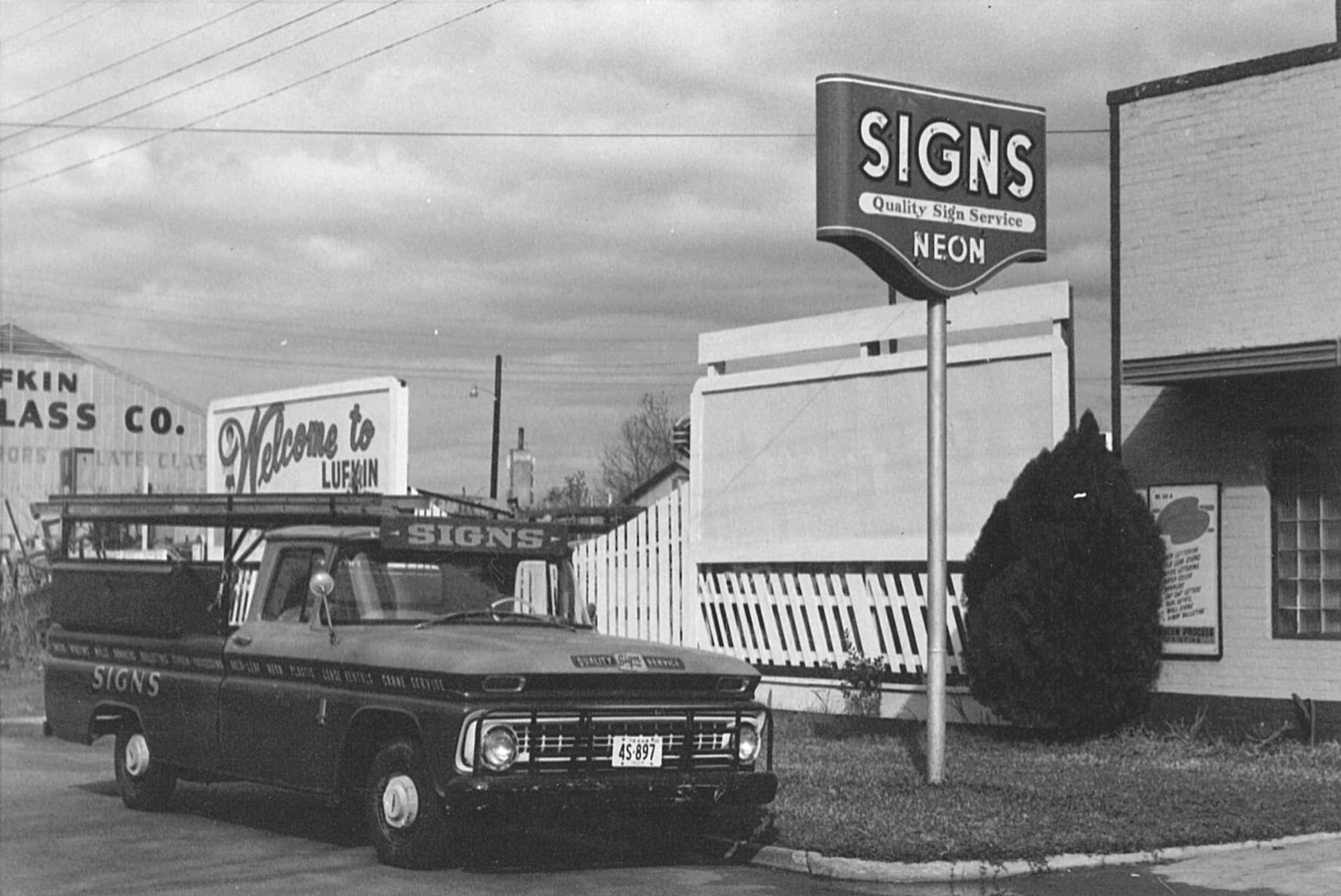 Truck parked outside of workshop premises and subtely adorned with signs that say "Signs". A sign mounted on a pole in front of the workshop says "Signs, Quality Sign Service, Neon".