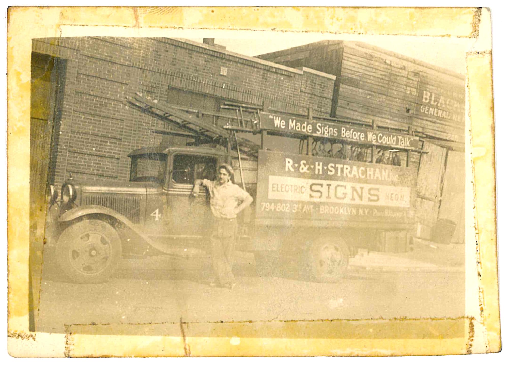 Man casually posing with his elbow leaning on the window ledge of his large truck. The vehicle is loaded with ladders and other sign painting equipment, and the lettering on its side reads "'We made signs before we could talk', R&H Strachan, Signs, Electric, Neon, Brooklyn, NY", along with phone contact information.