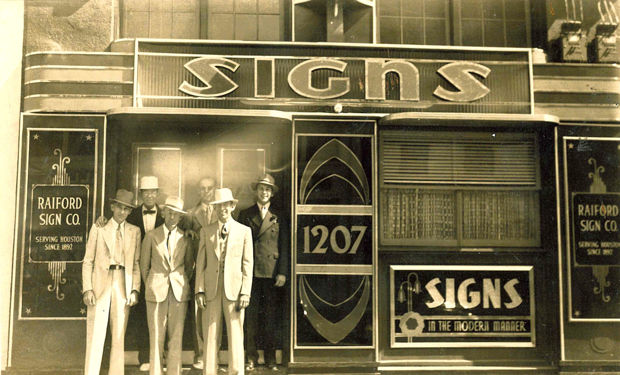 Six suited men posing in the doorway of a shop adorned with signs for the Raiford Sign Co.