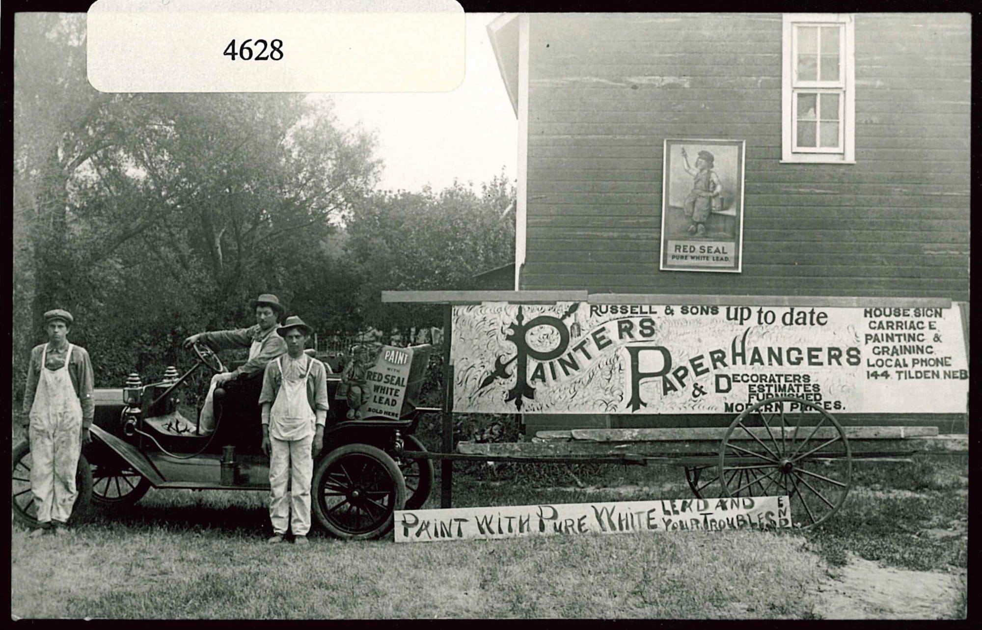 Men in overalls posing on/in front of an old car that's hitched to a large trailer adorned with a sign advertising their business that reads "Russell & Sons, Up-to-Date Painters, Paperhangers & Decorators" along with listings of other services.