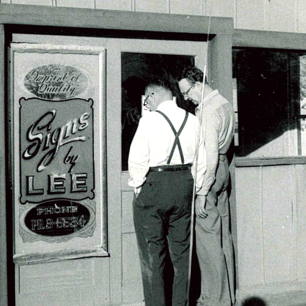 Two men with their backs to the viewer looking at a vertical sign mounted beside a doorway. The sign is a reverse glass piece in gold leaf and paint that reads "Signs by Lee, the Imprint of Quality, Phone PR.3-5534".