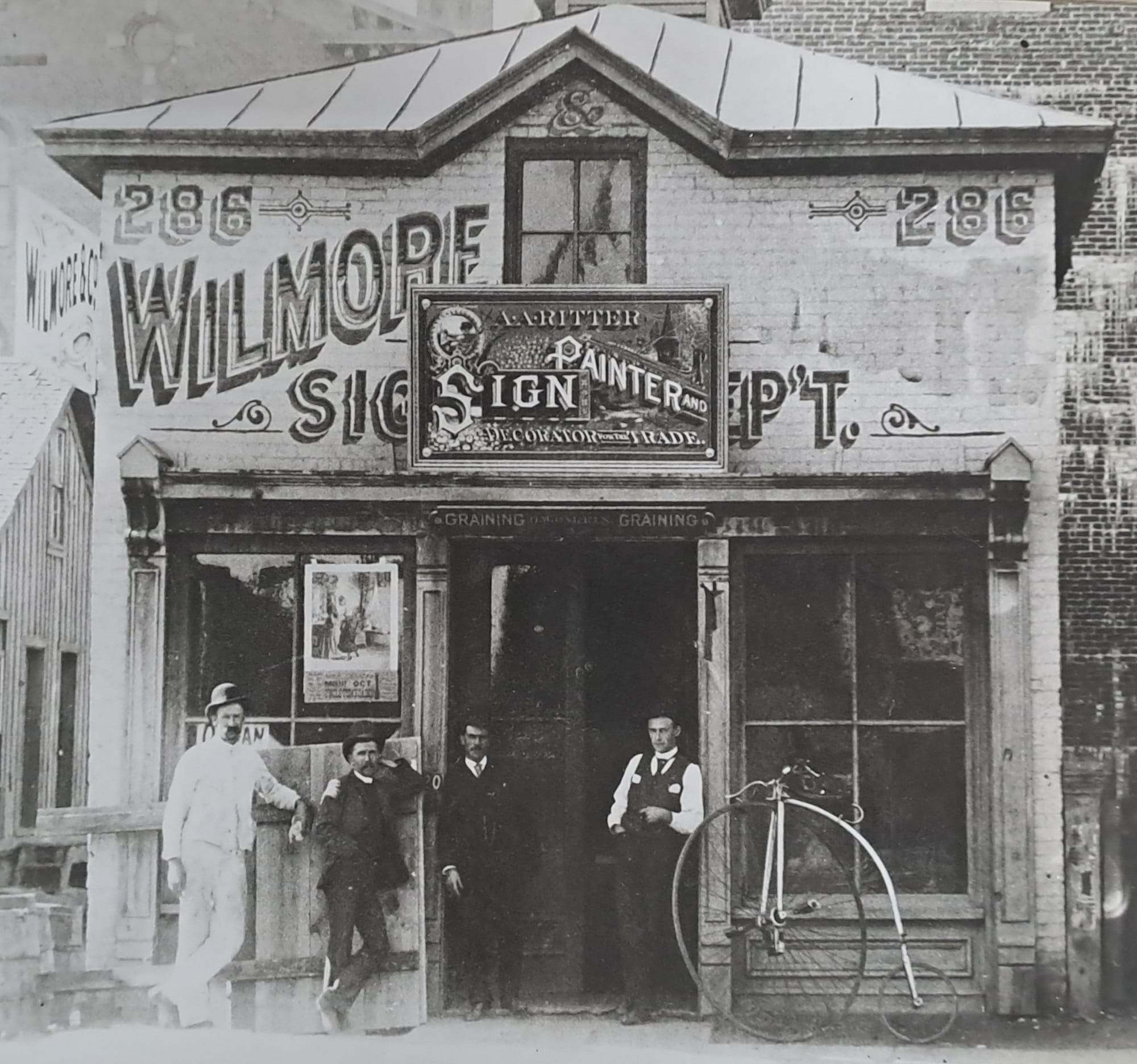 Group of men posing outside a shop that's adorned with a host of signs above the doorway and across the first floor. These advertise their sign business, and there is a penny-farthing style bicycle propped up outside too.