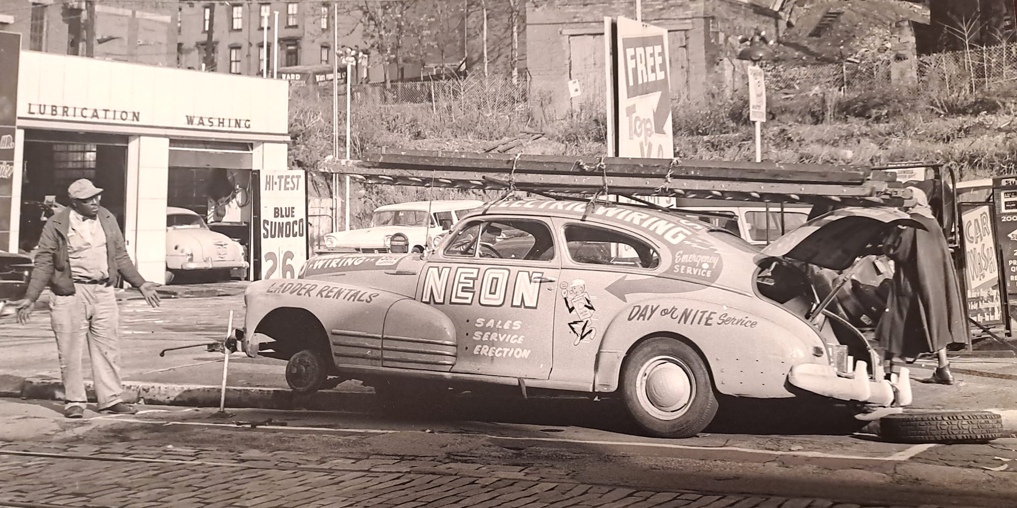 1940s style vehicle jacked up to change a tyre, and a man to the right looking at it with a despairing gesture. The car itself is lettered all over with signage for an un-named neon sign sales and repair business.