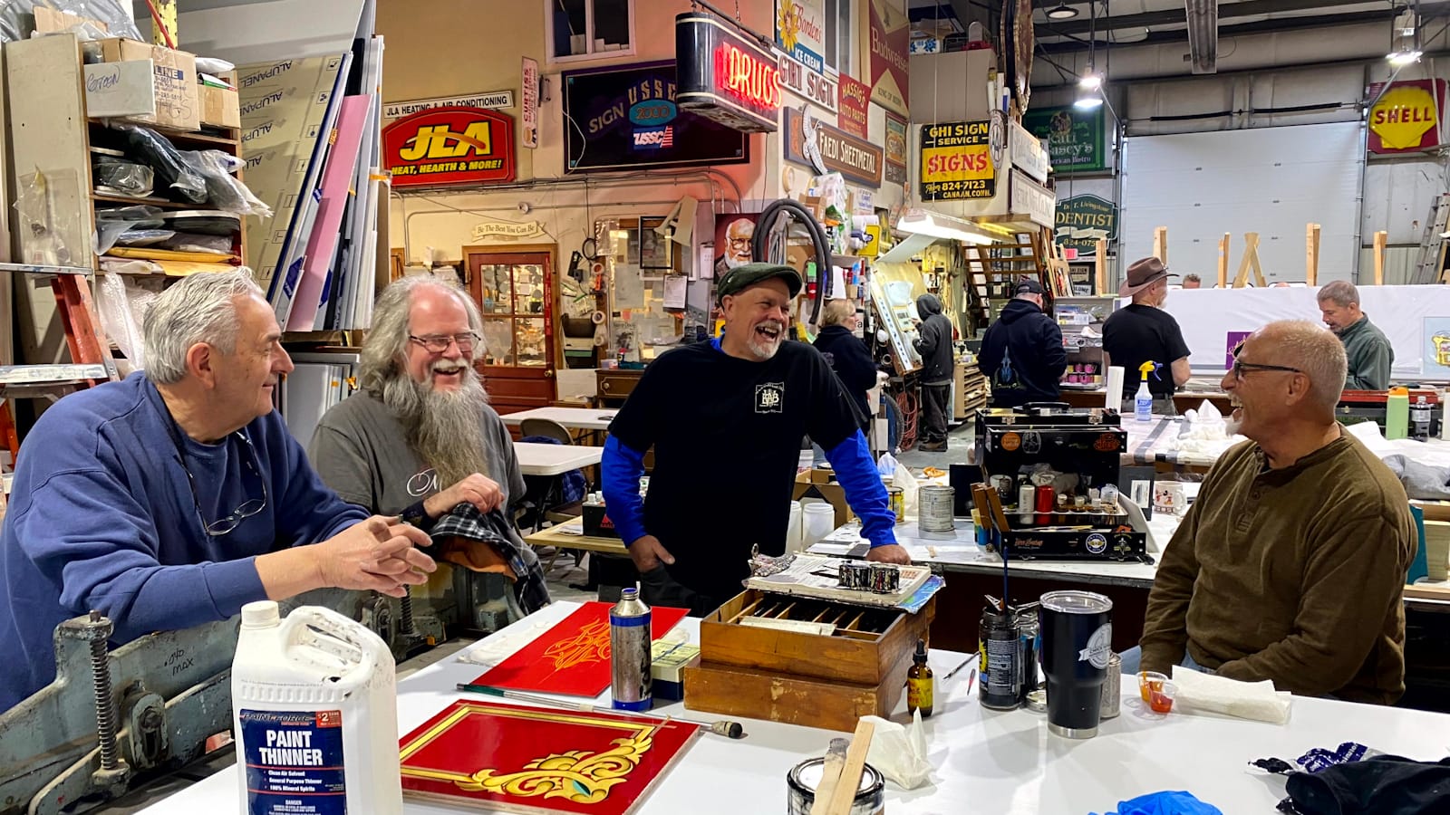 Scene in a sign shop adorned with dozens of signs. Four men are gathered around a table, smiling in conversation.