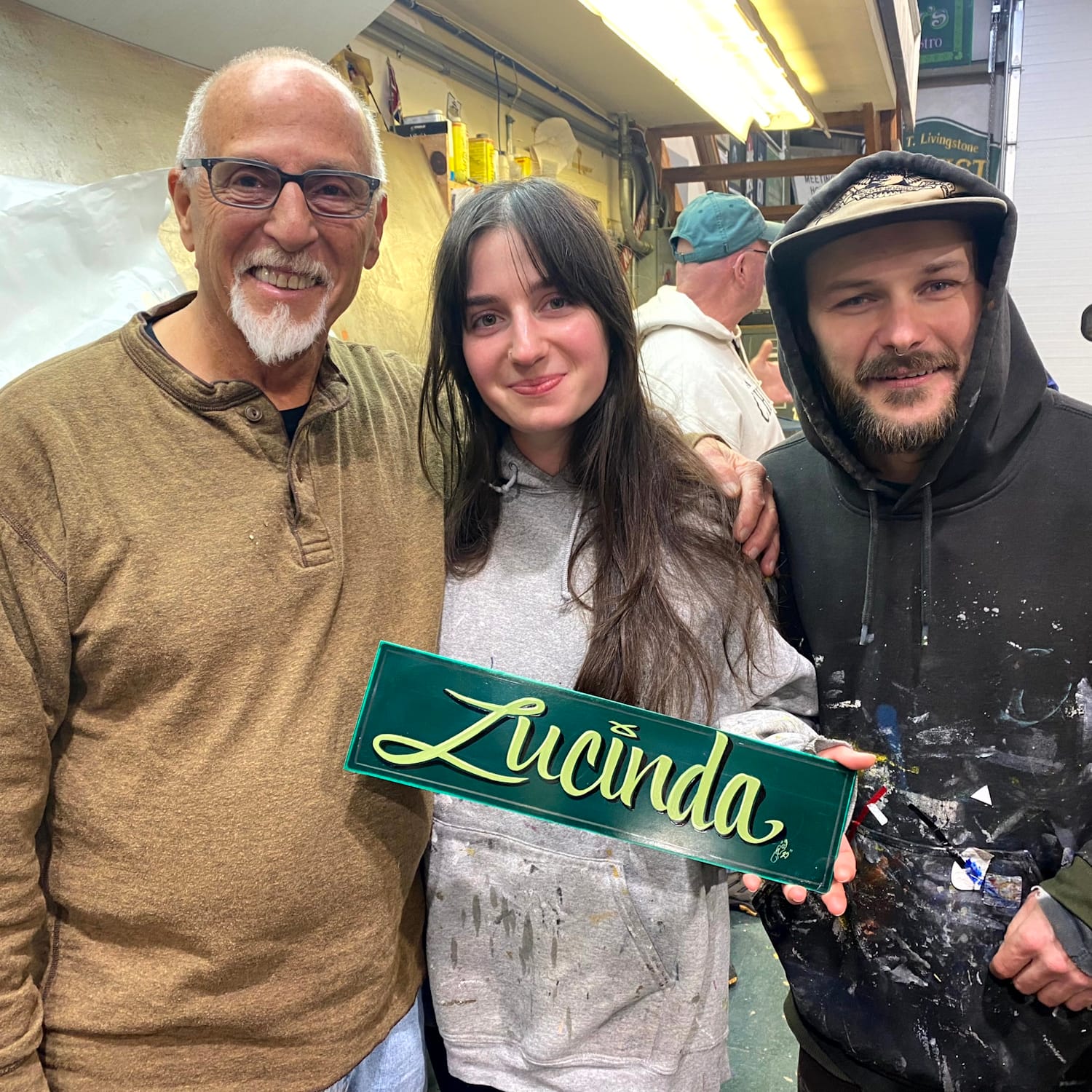 Three people smiling and posing for a portrait with the woman in the middle holding a green panel with script lettering that says "Lucinda".