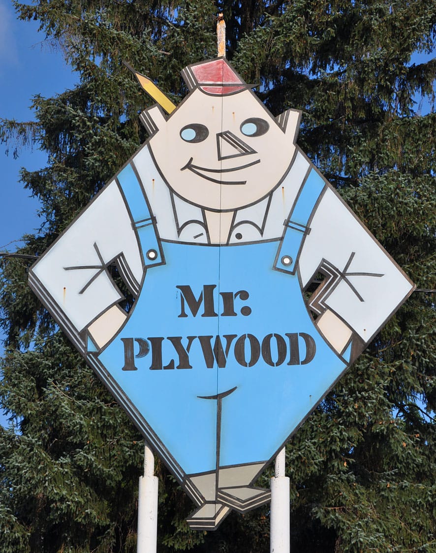 Large sign for Mr. Plywood, consisting of a cartoon character in an exaggerated diamond shape, wearing blue overalls, and with stencil lettering saying "Mr. Plywood" across the chest/stomach.