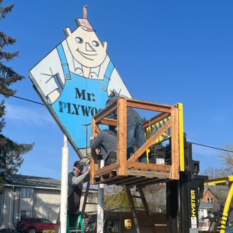 People on a ladder and fork-lift-elevated platform removing the Mr. Plywood sign from it two-pole mounting.