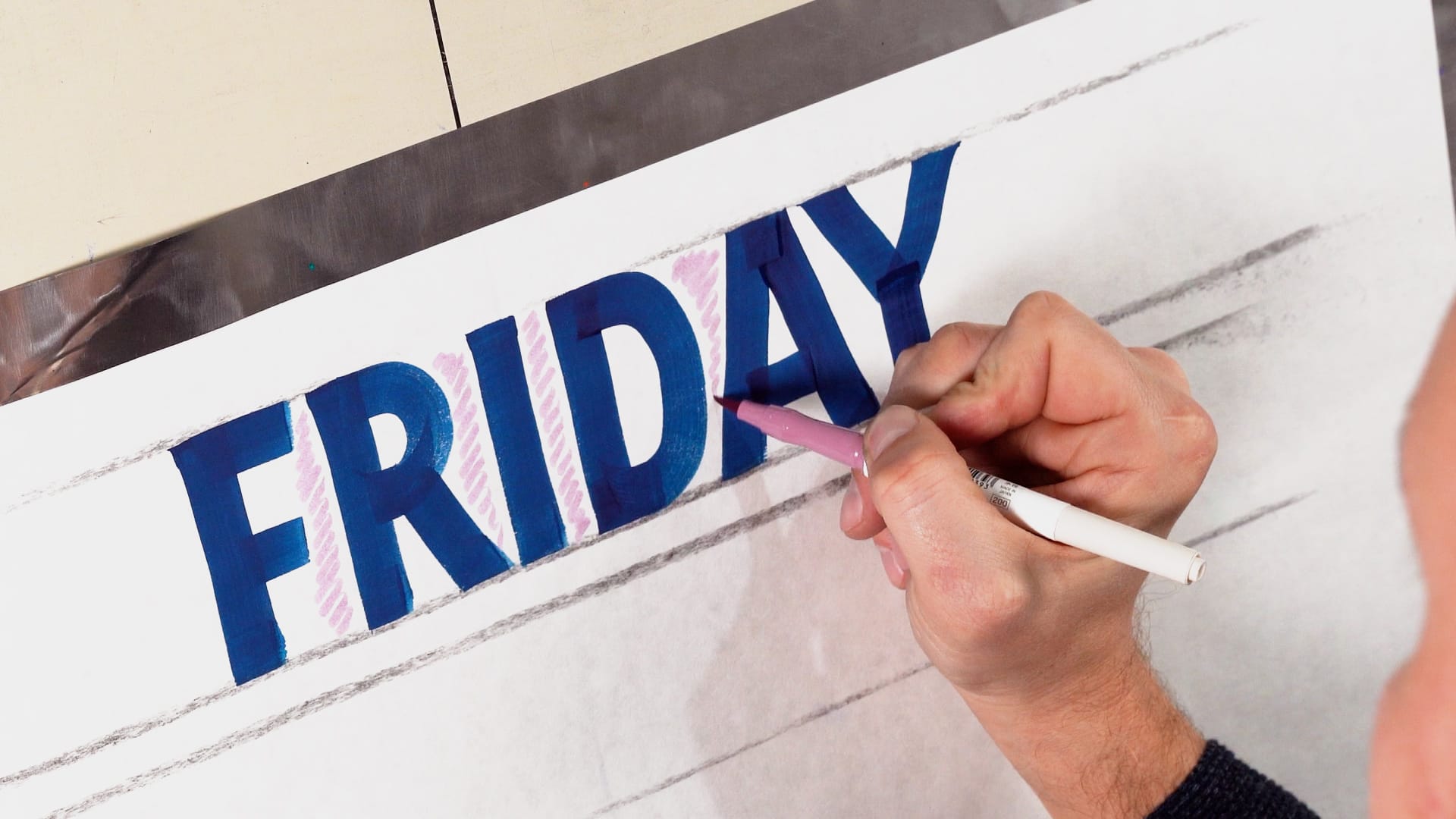 Photo of a hand lightly shading the negative spaces around block letters that have been painted in blue between horizontal guidelines.