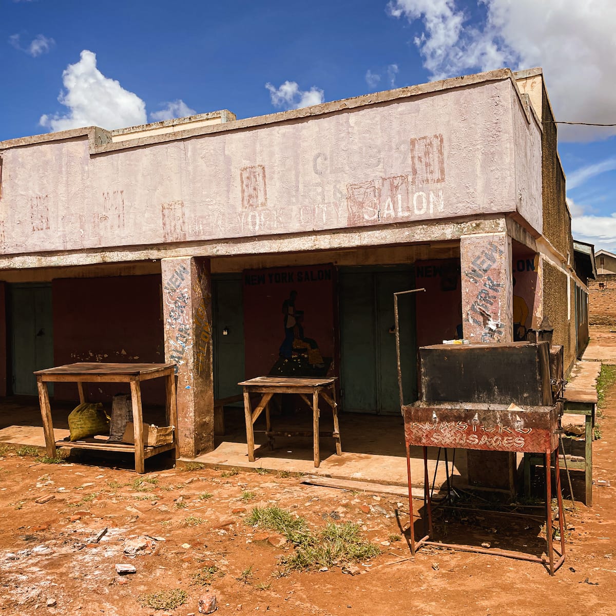 Dilapidated building with old tables and benches sitting outside. The frontage of the building forms an overhand to a shaded area underneath and has fading traces of painted signs on it.