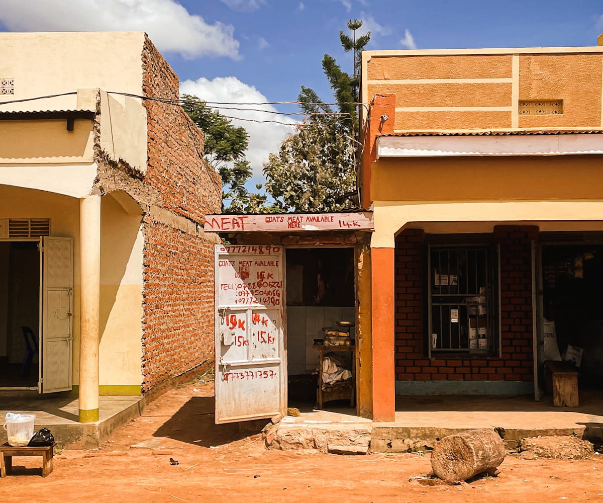 Lowrise buildings bathed in sunlight. In the middle is a small extension of one, protruding into an alleyway. Its white door is open and this, along with the lintel above, are scrawled with red paint advertising meat products and prices alongside phone numbers.