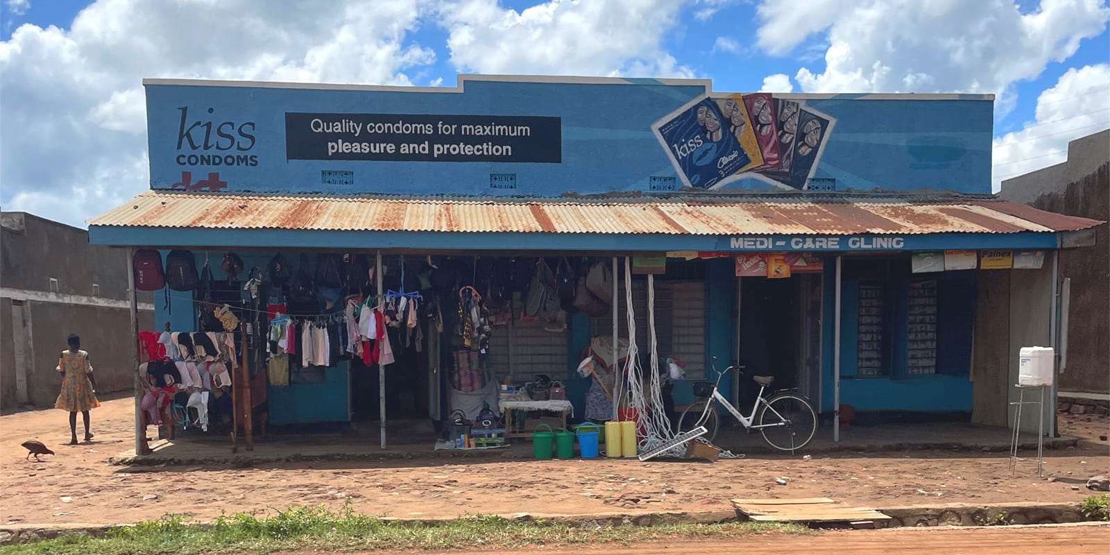 Low-rise shop building with a protruding canopy and clothes hanging from the this outside the premises that are located on the left of the photo. The premises on the right have the basic painted Medi-Care Clinic sign at the edge of the canopy, and above this is the large fascia sign advertising Kiss condoms. This is illustrated with a pictoria of five condom packets and a panel containing the slogan which is "Quality condoms for maximum pleasure and protection".