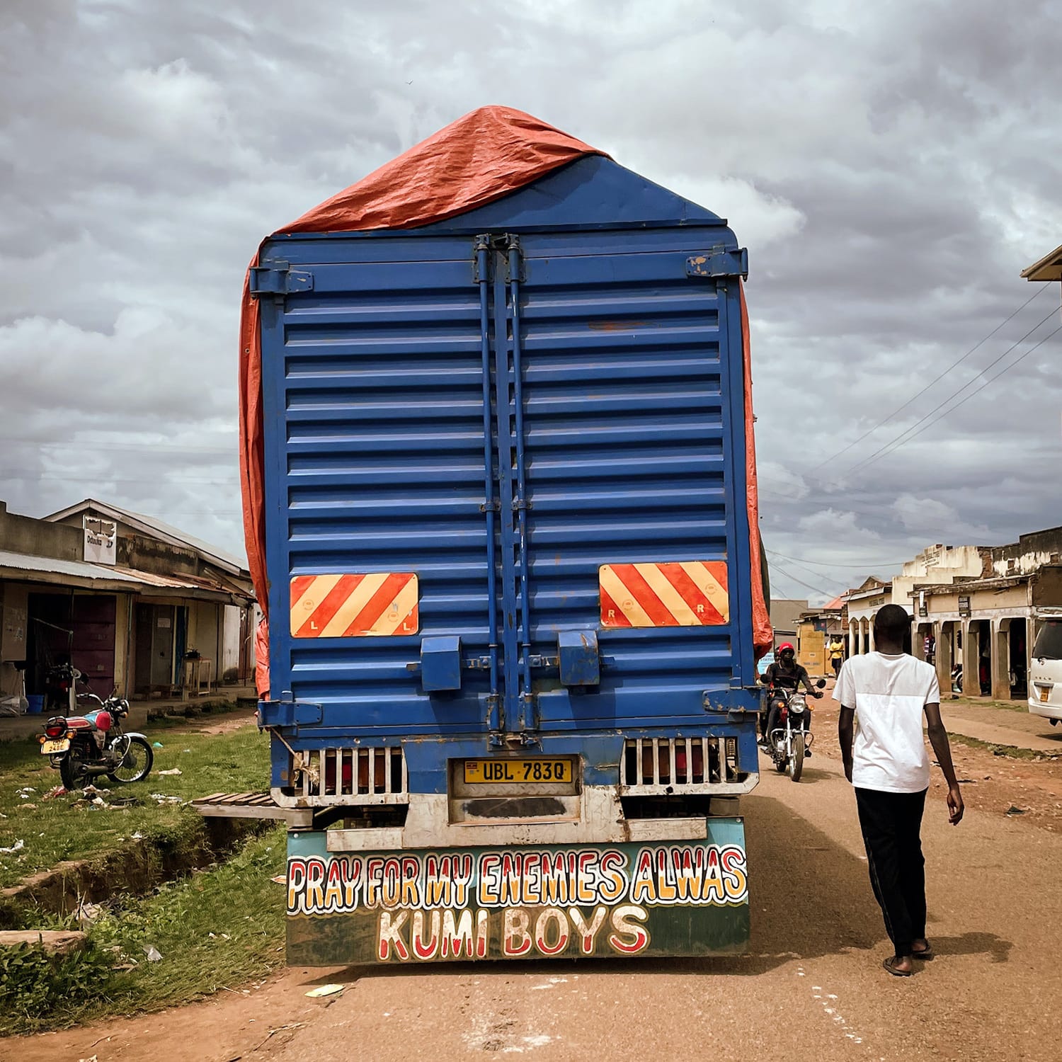 Rural African street scene with a large blue lorry blocking the view down the road. The painted panel hanging from the bottom of the back of the lorry is done in a crude bubbly lettering style with a blend from yellow to white to red going from the bottoms to the tops of the letters.