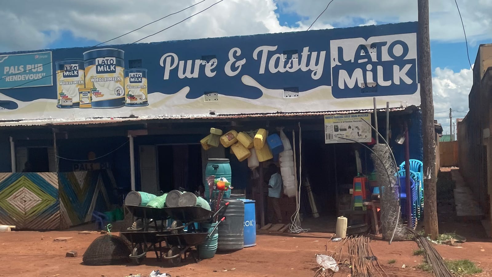 One-storey market building with various sizes and types of plastic containers and drums outside for sale. The fascia panel has a painted sign advertising 'Pure & Tasty Lato Milk' which includes a pictorial of different cans of their milk products.