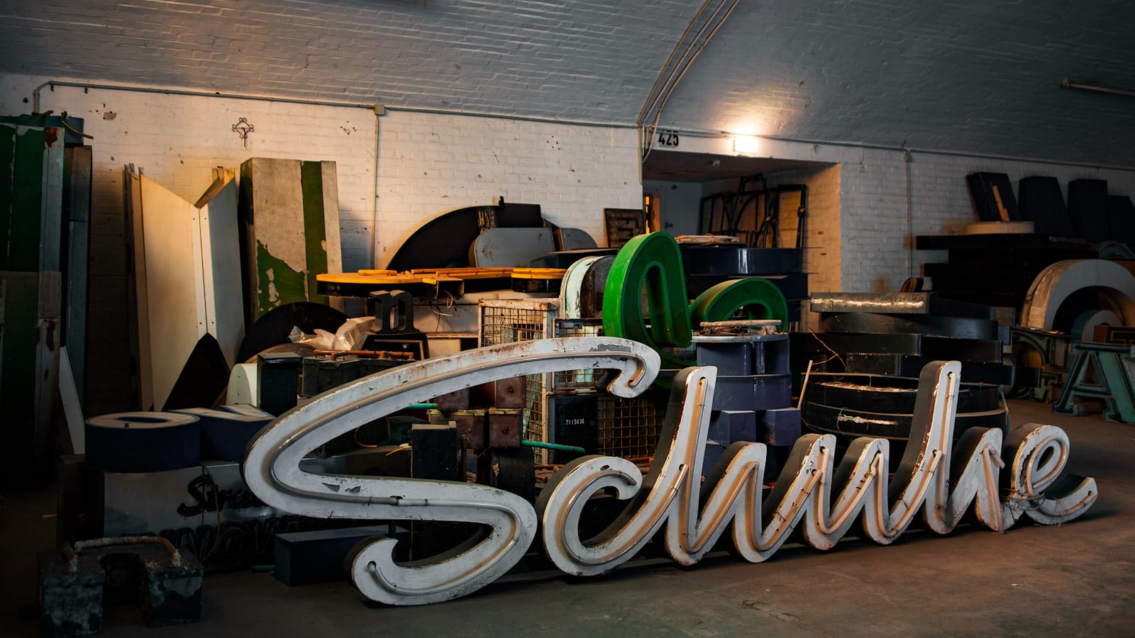 Script channel letters in the foreground with assorted letters and objects piled up behind them in an arched warehouse space.