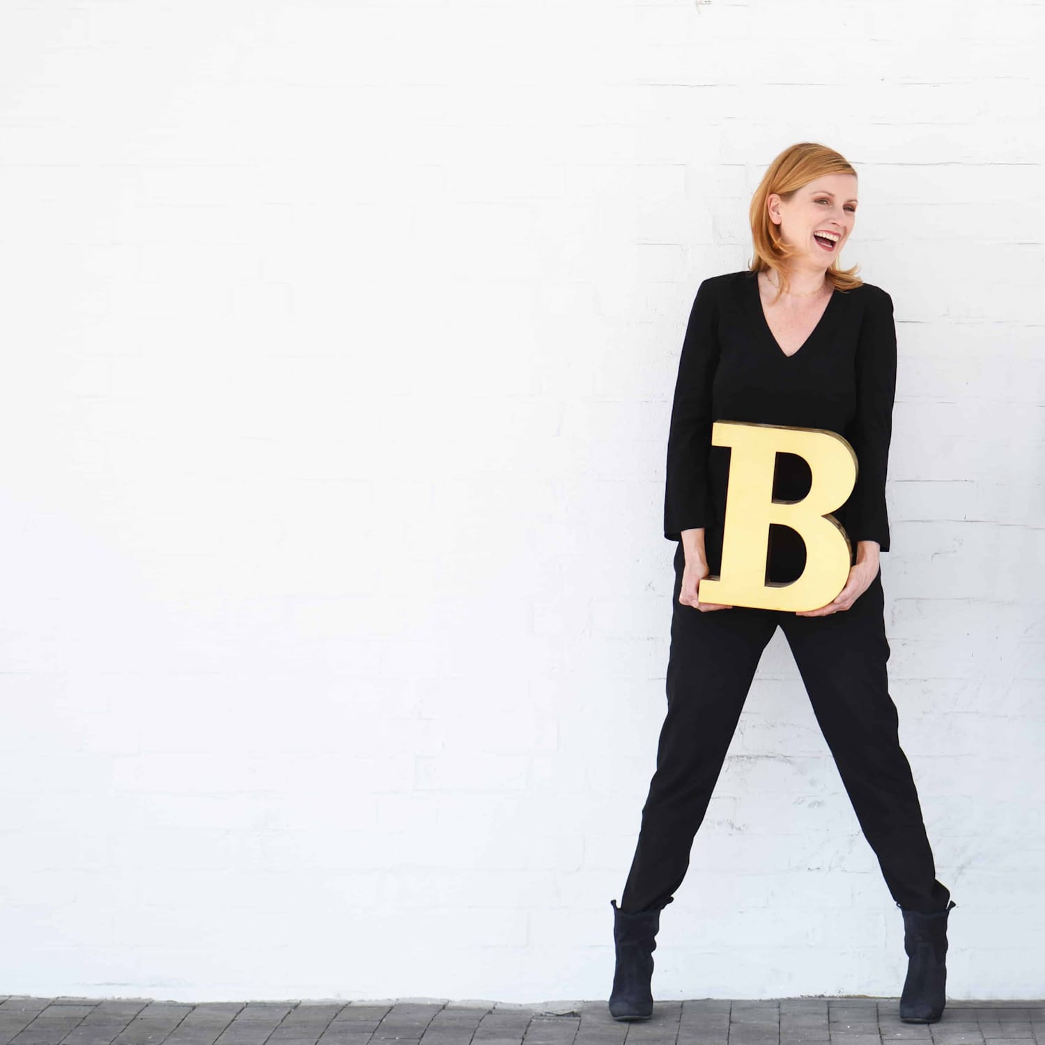 Woman dressed in black posing in front of a white wall holding a golden letter B.