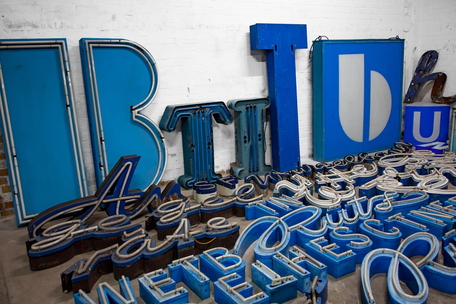Channel, neon, and other letterforms making use of blue in their colour schemes arranged on the floor and against a wall.