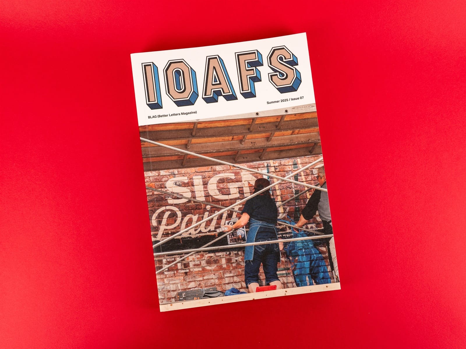 Magazine cover with masthead IOAFS set above a roughly square picture of two people on a scaffold painting lettering onto a brick wall.