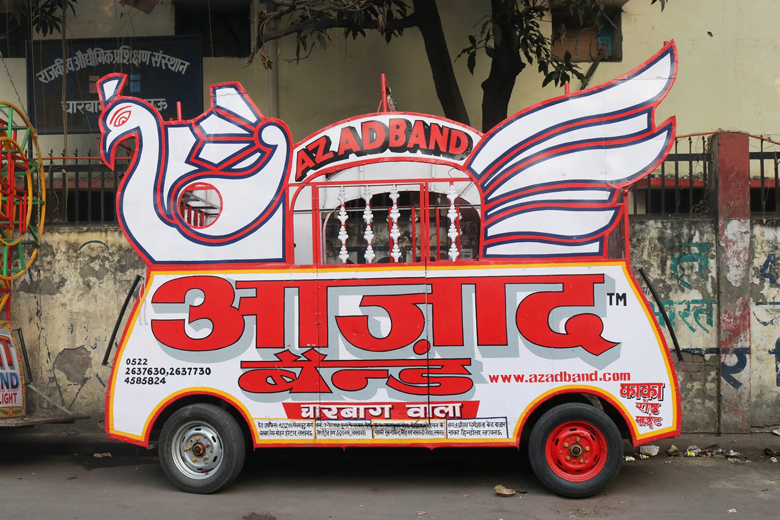 A motorised trolley for a brass band standing on the street. The trolley features a large peacock motif on the top with the name “Azad Band”, in English, incorporated within the illustration. On the bottom, the name repeats in Devanagari, along with information about the business. 