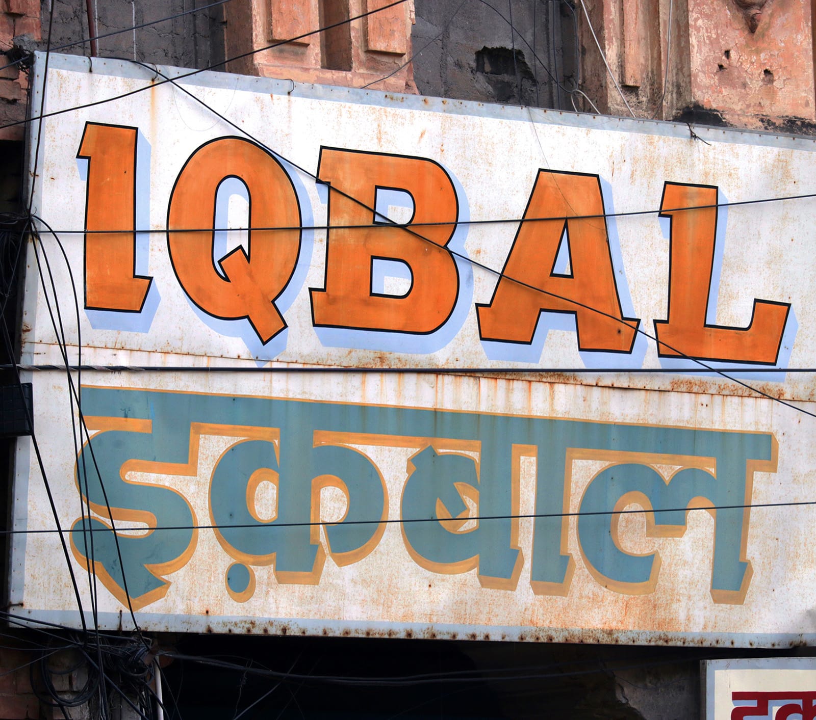 A white sign with a thin blue border that reads “Iqbal” in Latin and Devanagari. The Devanagari lettering copies the serifs of the Latin text.
