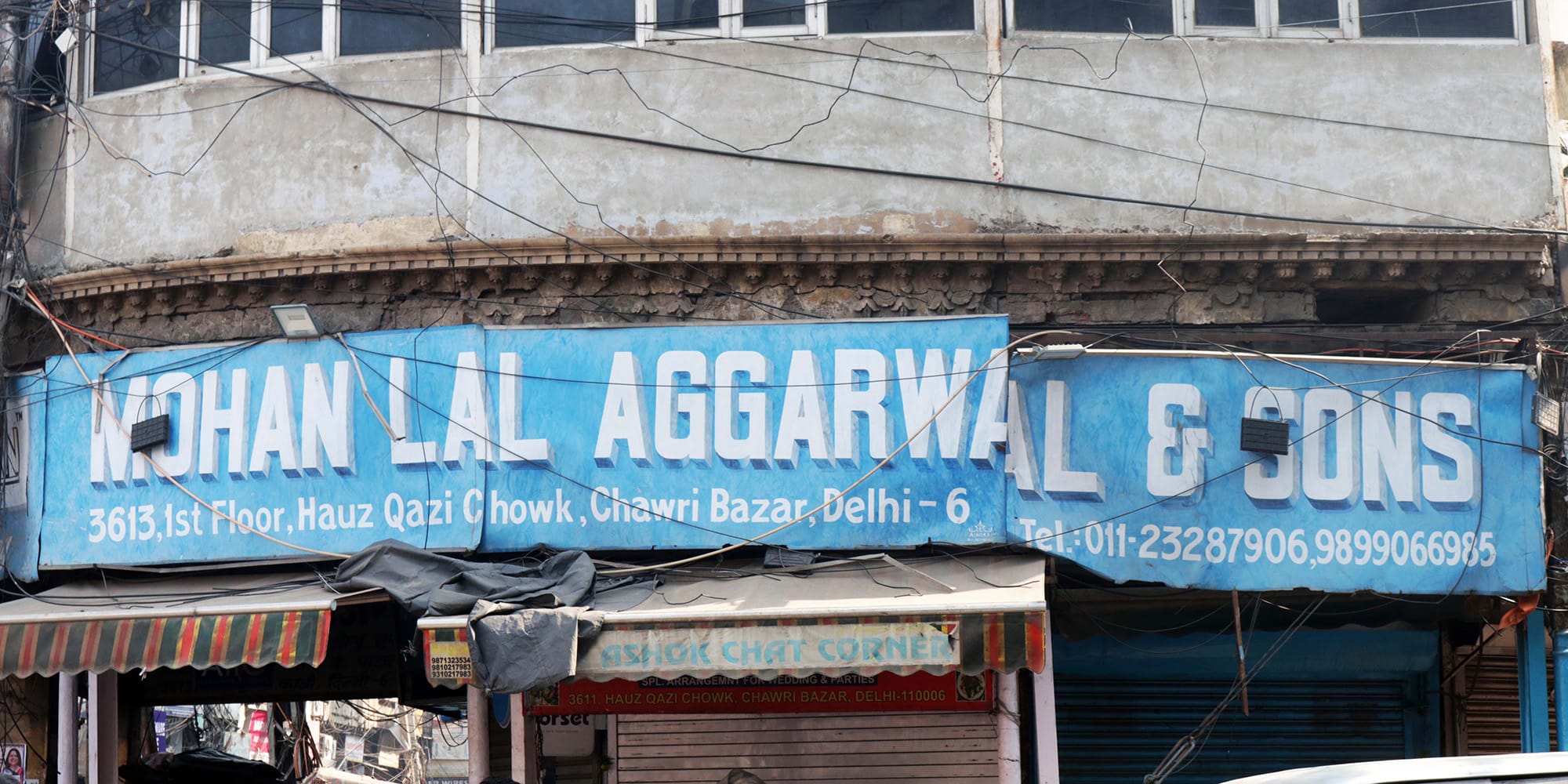 The front of a gray building with large windows and a wide blue sign that reads “Mohanlal Agarwal & Sons”, along with the building address and business phone numbers.