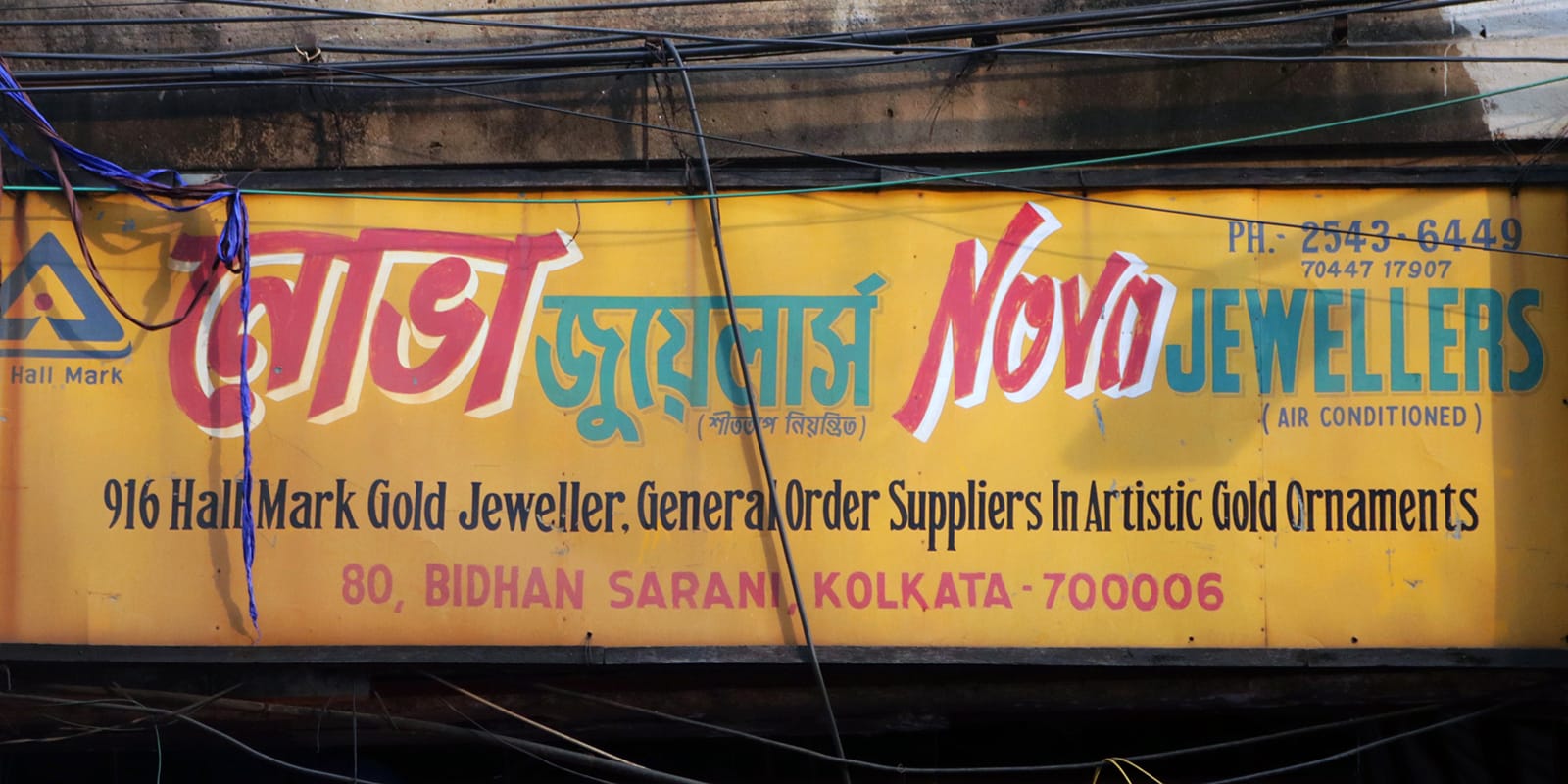 An orange sign on a decrepit brown building. The sign reads “Nova Jewellers” in Bengali and Latin, and mentions the shop’s services and address.