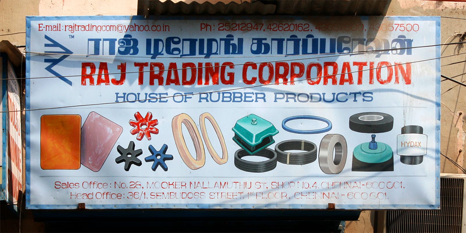 Multiscript sign for Raj Trading Corporation featuring Tamil text in blue and Latin text in red, as well as illustrations of rubber products the shop sells.