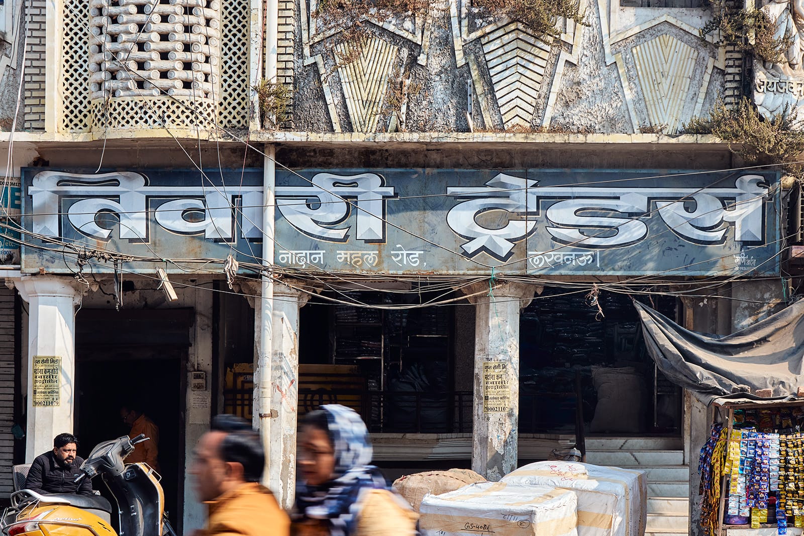 The façade of a white Art Deco building with large boxes on its steps, and a couple passing in front of it on a two-wheeler. The building has a sign in greyish blue, which reads “Tiwari Traders” in Devanagari.