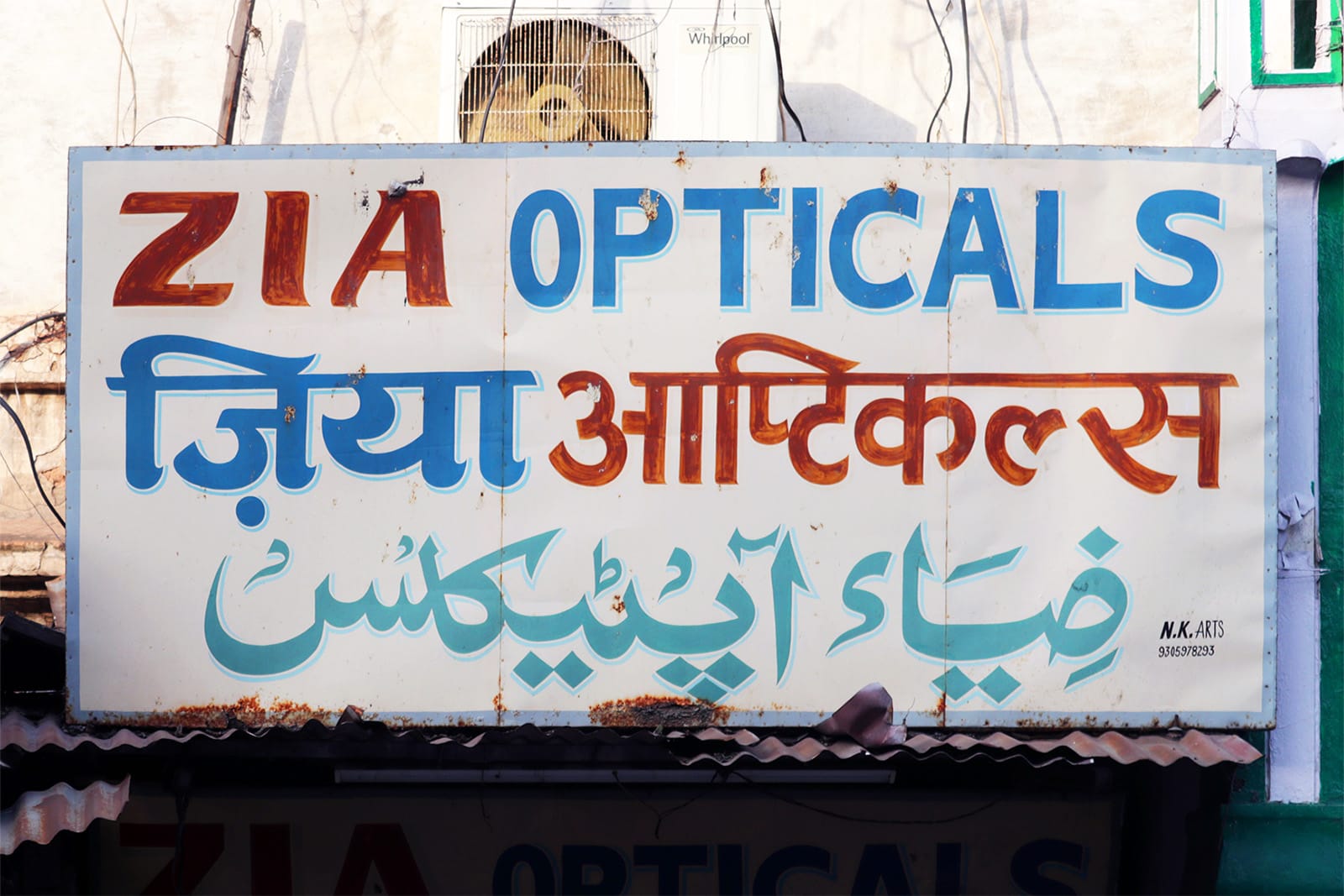 A white sign with a thin blue border, which reads “Zia Opticals” in Latin, Devanagari and Nastaliq in successive lines.