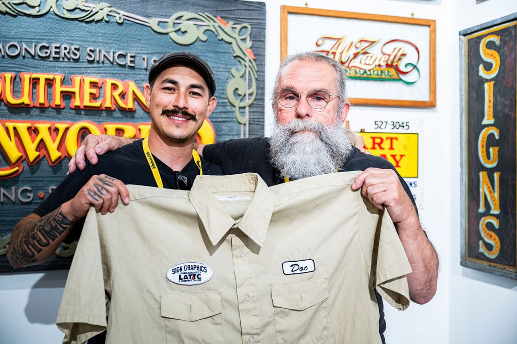 Two men posing for the camera in front of some hand-painted signs. They are holding a large beige shirt that has patches sewn onto it left and right and which read "Sign Graphics, LATTC" and "Doc" respectively.