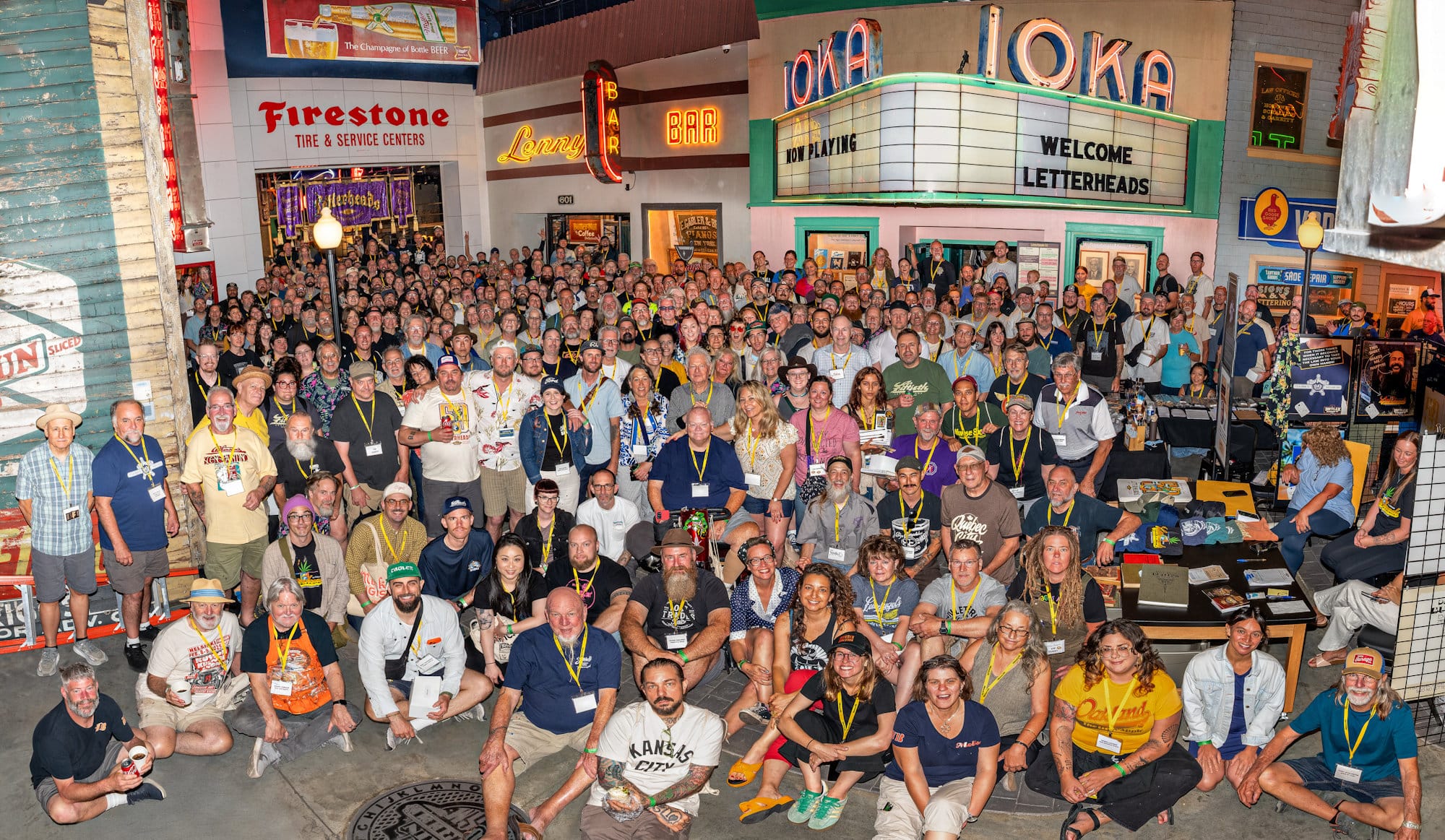 Group photo with hundreds of people in it. They are in a museum/gallery setting which is filled with old signs, the most prominent of which is the IOKA theatre marquee with its neon letters on top and the readerboard below that says "Welcome Letterheads".