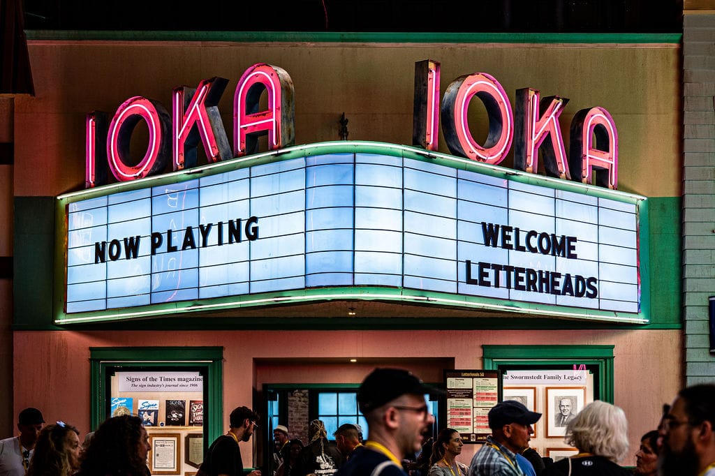 Illuminated theatre marquee sign with neon letters above that read "IOKA". The readerboard then says "Now Playing" (left) and "Welcome Letterheads" (right) and below there are people's heads visible as they gather in small groups and talk.