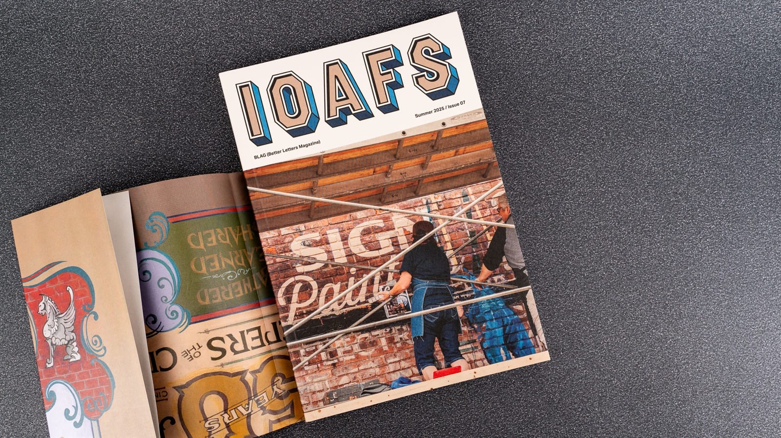 Magazine cover with IOAFS as the masthead and a picture below that showing people on a scaffold painting a sign. There is a cover wrap that is partially removed and this has hints of colourful lettering visible.
