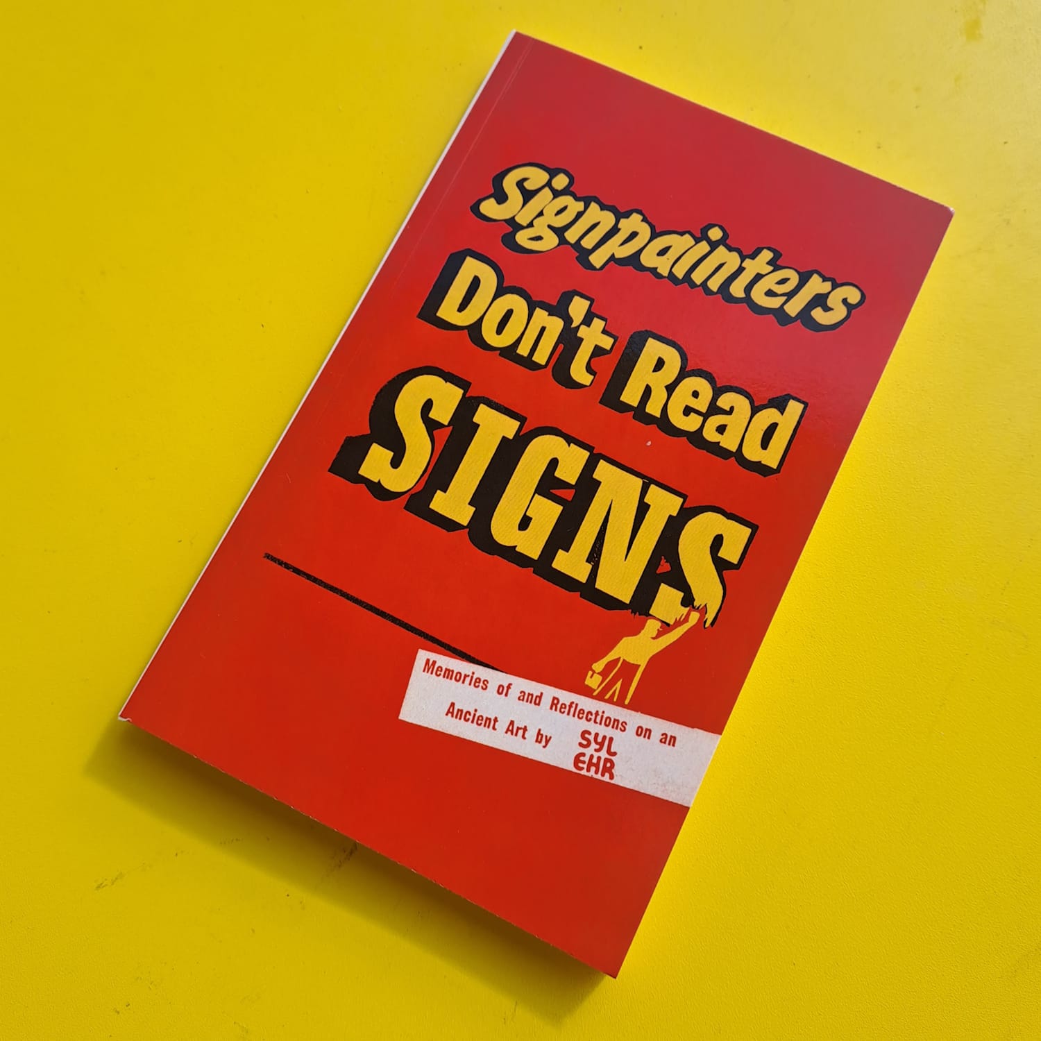 Photo of the book's cover which has the title set in yellow lettering in sign-painted lettering with a small illustration of a painter finishing the last S of Signs at the bottom. A white band cuts in from the right of the cover with smaller red lettering that reads "Memories of and Reflections on an Ancient Art by Syl Ehr".