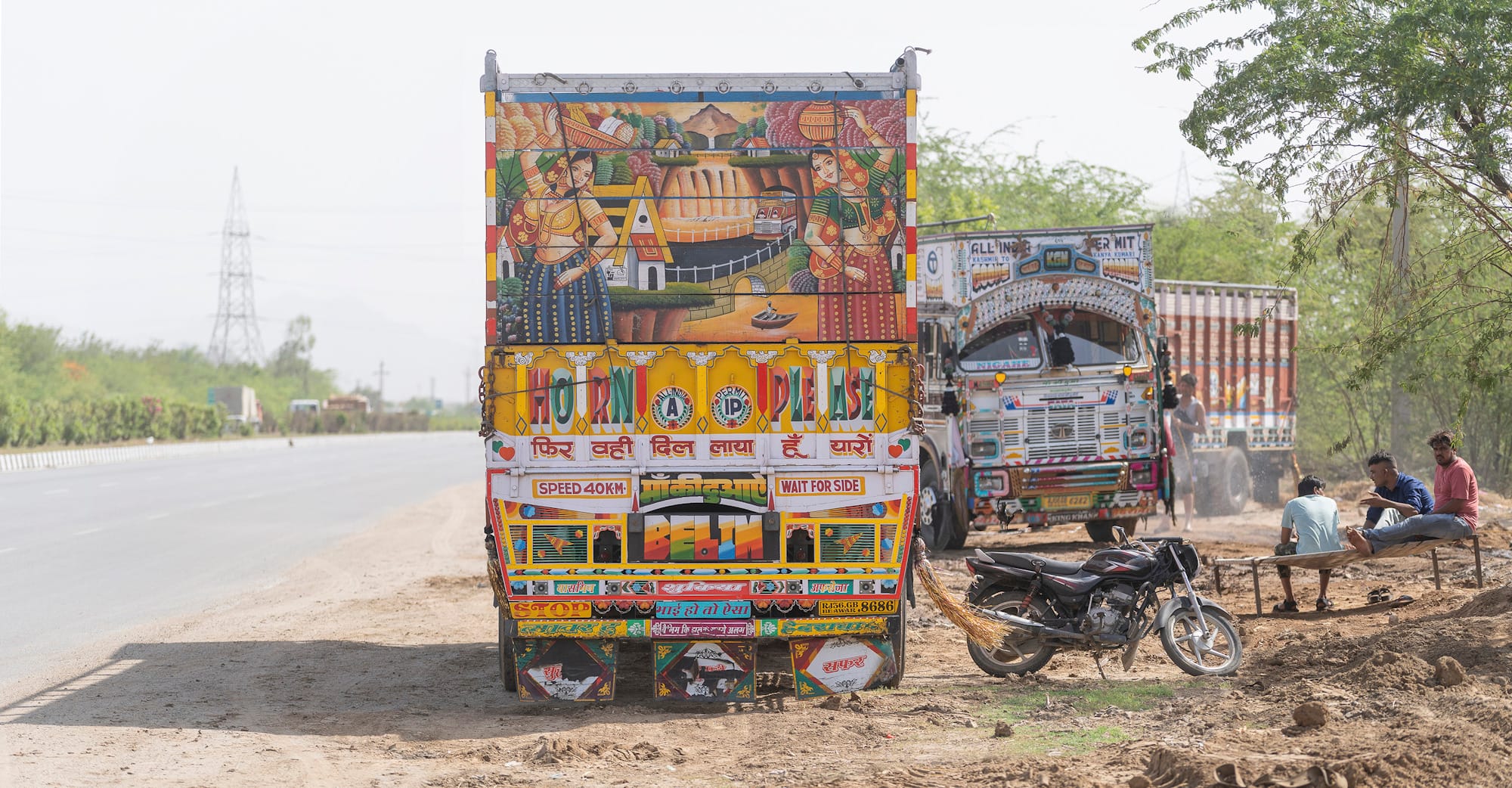 Three heavily decorated trucks, one more prominent in the foreground, and a motorbike parked at the side of an empty road with three men sat on simple benches nearby.
