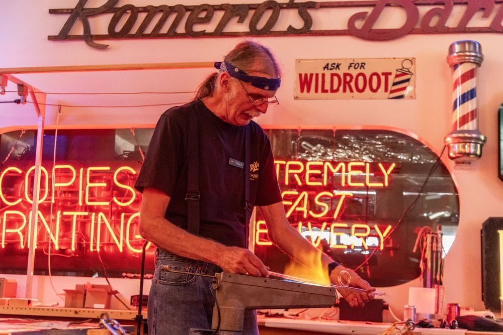 Man holding a glass tube over a flame with various signs from the museum's exhibits as the backdrop.
